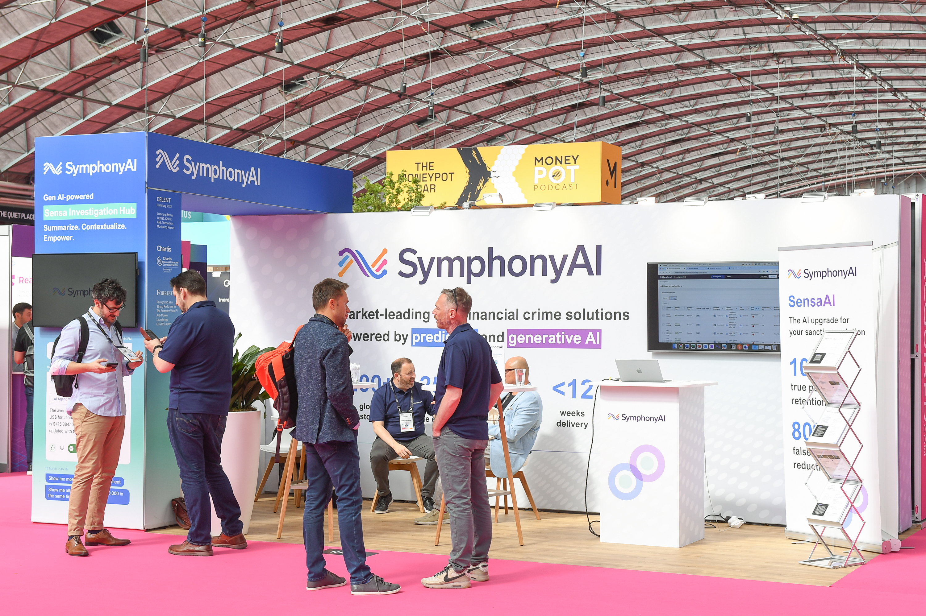 Photo of an 
exhibition stand with three open sides. There is a back wall with a graphics showing a white background and company logo and a screen on the right hand side. There is a branded counter on the right centre side of the stand next to a steel literature rack displaying brochures. On the left centre there is a table and chairs with people sat down having a conversation. There is rectangular arch on the 
far left of the stand attached to the back wall displaying blue branded graphics. There are people stood having a conversation in front of the stand.